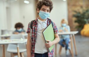 portrait of cheerful little school boy wearing mask to prevent the spread of covid19 looking at camera, holding textbook while posing in a classroom