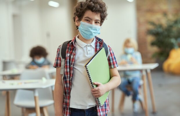 portrait of cheerful little school boy wearing mask to prevent the spread of covid19 looking at camera, holding textbook while posing in a classroom