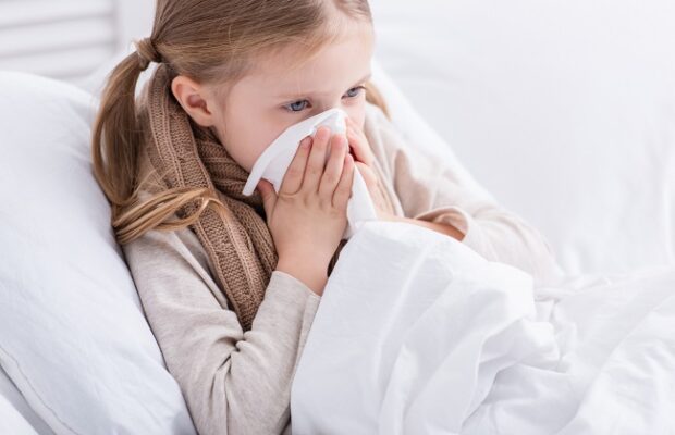 sick child with scarf over neck lying in bed and blowing nose in tissue at home