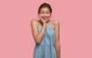 joyful pretty dark haired woman has toothy smile, keeps hand on cheek, dressed in denim dress, models against pink background, happy to recieve compliment from husband, expresses positiveness