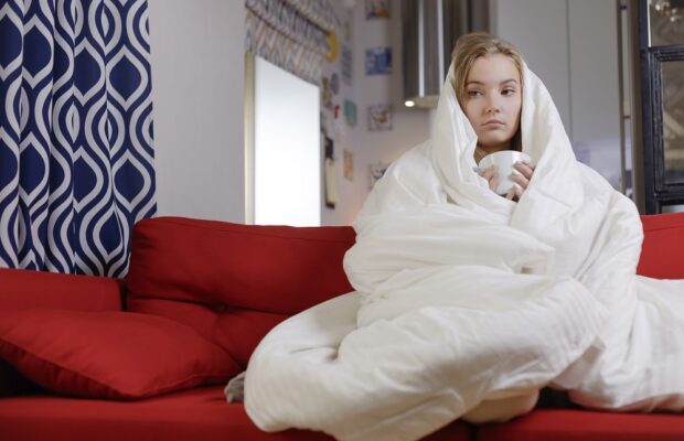 woman with mug wrapped under the covers on couch cold weather, having a cold and lonely mood