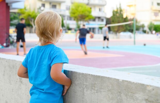 a little three year old boy in a blue t shirt watches teenagers playing with a ball on an outdoor sports field, dreaming of playing sports